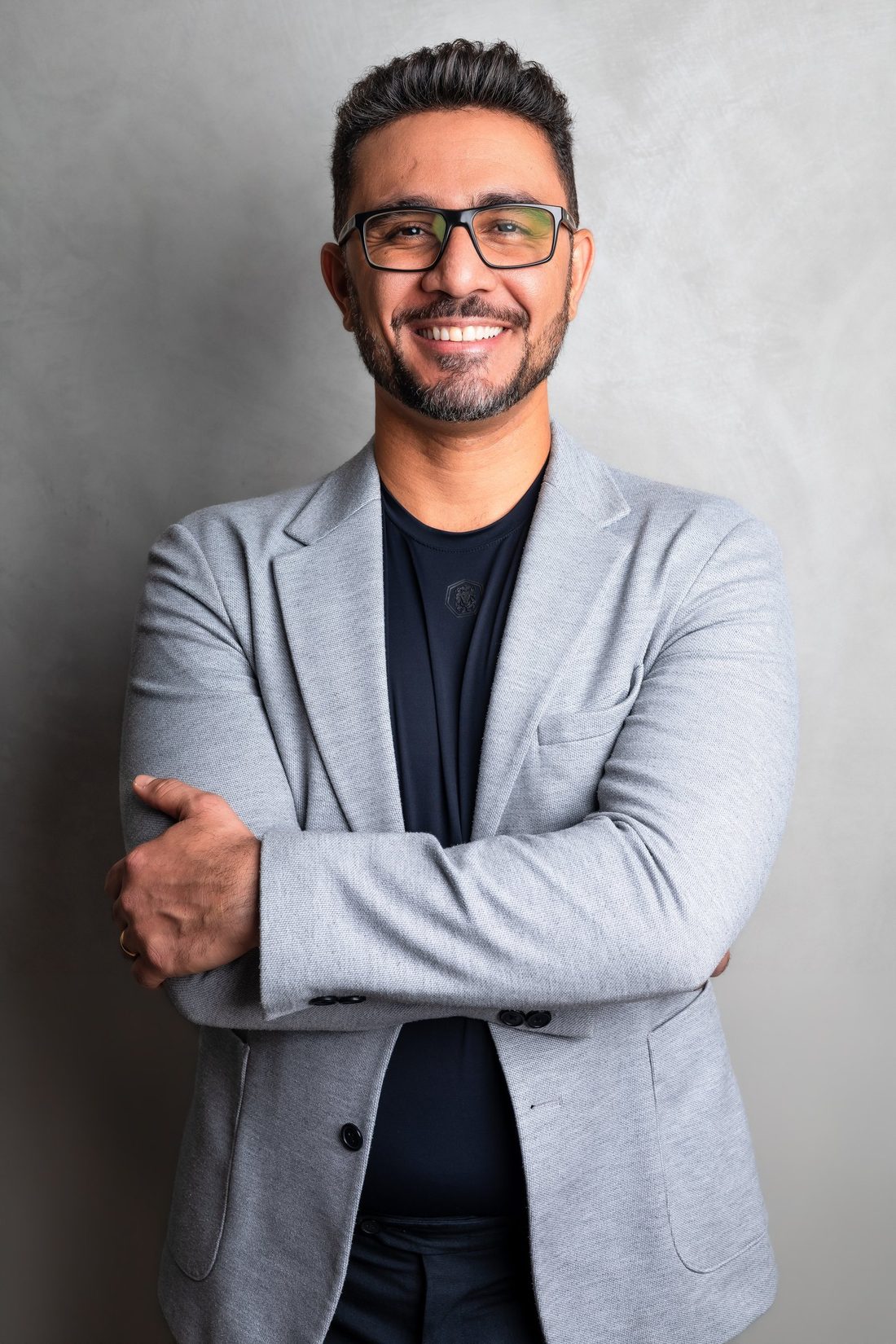 Adeilton Rufino de blazer cinza, sorrindo, de braços cruzados